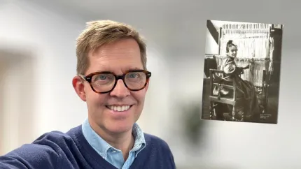En leende man med glasögon tar en selfie inomhus, med ett svartvitt foto av en vävstol på väggen..