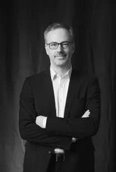 Svartvit studiofotografering av en man i kostym och glasögon med armarna i kors mot mörk bakgrund..