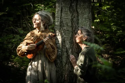 Två kvinnor vid ett stort träd i skogen; en håller en fiol medan den andra lutar sig mot stammen i solglimtar.