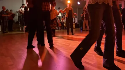 Människor dansar på ett trägolv inomhus med röd scenbelysning, medan musiker och publik syns i bakgrunden..