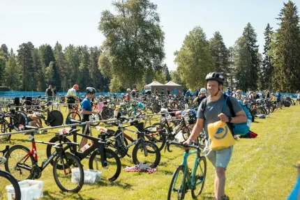 Cyklar står uppradade i ett växlingsområde på en gräsmatta, med deltagare och träd i bakgrunden..