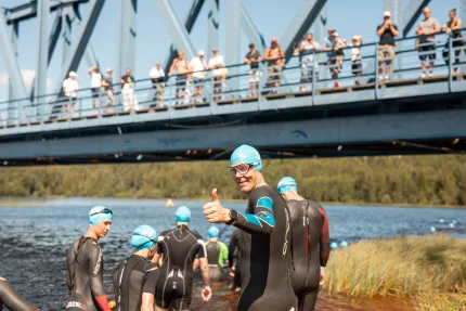 Triathleter i våtdräkter och blå badmössor står vid en sjö; en man ger tummen upp medan publik ser på från bro..