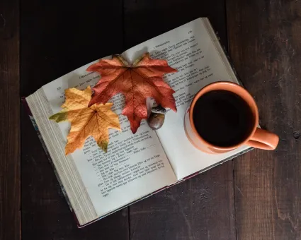 Uppslagen bok med två höstlöv och ett ekollon på sidorna, bredvid en kaffekopp på mörkt träbord..