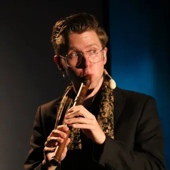 En man i mörk kostym och scarf spelar ett träblåsinstrument på scen, med glasögon och headsetmikrofon..