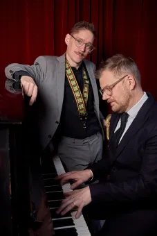 Två män i kostym vid ett piano; en står och lutar sig, den andra sitter och spelar framför röda draperier..