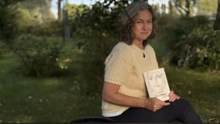En kvinna sitter utomhus i en grön park och håller upp en bok med ett ansikte på omslaget..