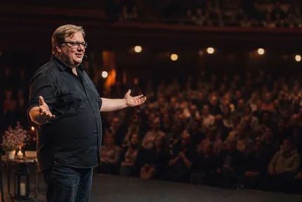 Man med glasögon står på en scen med utsträckta armar och talar inför en stor publik i en teater..