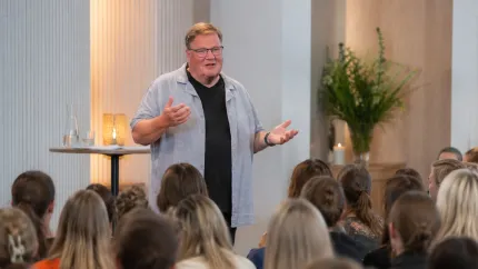 En man med glasögon gestikulerar och talar till en sittande publik i en ljus inomhuslokal med växter..