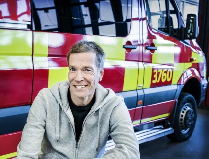 En leende man i grå huvtröja sitter framför ett rödgult räddningsfordon med siffror på sidan, inomhus..