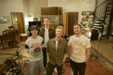 Fyra män står i en musikstudio med mikrofonstativ, kablar, mattor och instrument runt omkring..