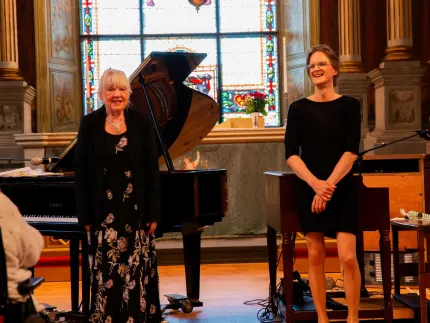 Två kvinnor står framför en flygel i en kyrka med stora färgade glasfönster, mikrofoner och blomsterarrangemang..