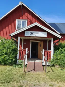 Röd träbyggnad med vit veranda och ramp, öppen dörr och en skylt ovanför entrén under blå himmel..
