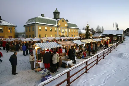 Snötäckt torg med marknadsstånd och människor framför en gul byggnad, julgran och ljusslingor i skymning..