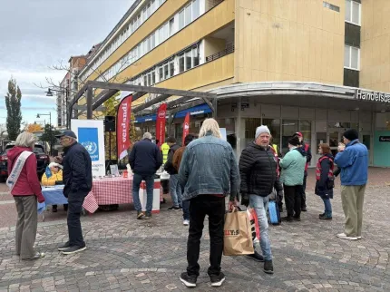 Människor står på stenlagt torg vid bord med duk och flaggor utanför en gul byggnad en mulen dag..