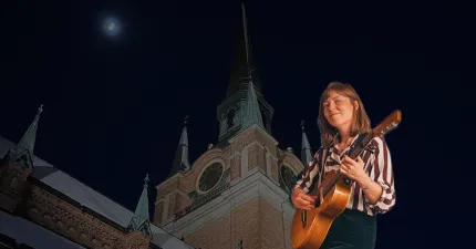 En person håller en akustisk gitarr framför ett upplyst kyrktorn med klockor mot mörk natthimmel och måne..