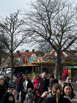 Folk samlas vid en färgglad karusell på en marknad, med bara trädgrenar och grå himmel i bakgrunden.