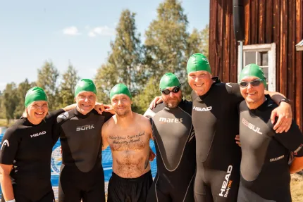 Sex personer står tätt ihop utomhus, fem i svarta våtdräkter och gröna simmössor, vid en blå pool..