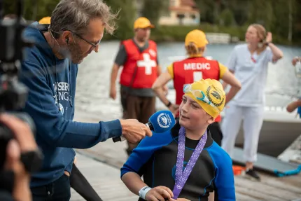 En pojke i våtdräkt och badmössa intervjuas med mikrofon på en brygga, med sjukvårdspersonal vid vattnet..