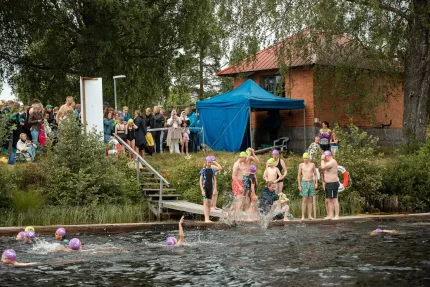 Grupp barn och vuxna med färgglada badmössor vid en brygga, medan flera simmar i mörkt vatten nära land..