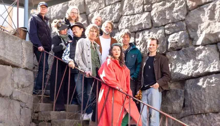 Grupp med vuxna personer står på en utomhustrappa vid en stor stenmur, i dagsljus med jackor och hattar..