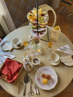Runt bord dukat för afternoon tea med kakfat, frukt, smörgåsar, tekoppar, glas och liten vas med blommor..