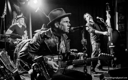 Svartvit foto av band på scen: sångare med hatt och gitarr vid mikrofon, trummis och ståbasist i bakgrunden..