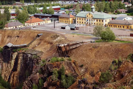 Vy över gruvschakt med branta slänter och gångbro, framför en gul stationslik byggnad och andra hus..