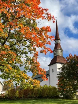 Vit kyrka med högt torn och klocka, inramad av träd med röda och gula höstlöv mot blå himmel..