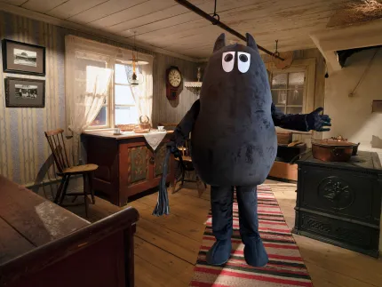 Stor mörk muminkostym står på en randig trasmatta i ett rustikt kök med vedspis, bord och fönster..