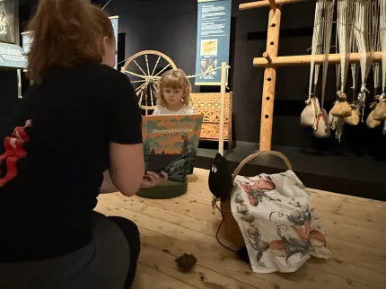 En vuxen läser en bok för ett barn i en museimiljö med spinnrock, träram, rep och vävredskap i bakgrunden..
