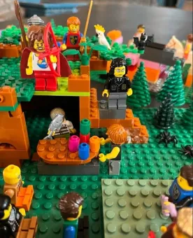 Färgglad LEGO-scen med flera minifigurer på grön basplatta, träd och en liten grotta med detaljer.