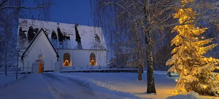 En vit kyrka med snötäckt tak i skymning, varma ljus i fönster och en gran med julbelysning till höger..