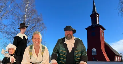 Fyra personer i historiska kläder framför en röd kyrka med högt torn, barträd och klarblå himmel..