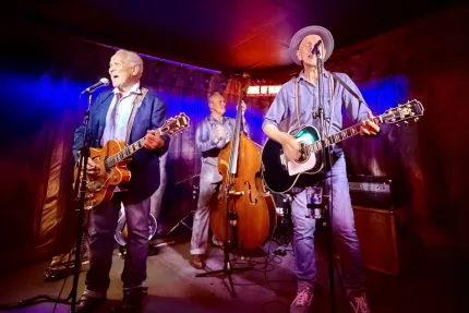Tre musiker uppträder på en liten scen med mikrofoner, två gitarrer och en kontrabas i färgat ljus..