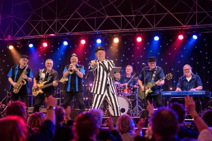 Musikband uppträder på scen med sångare i randig kostym, blå och röda scenljus samt publik i förgrunden..