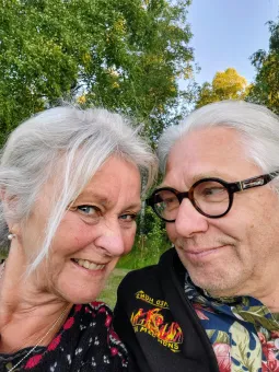 Närbild av två leende äldre personer utomhus med grönskande träd i bakgrunden och klar himmel..