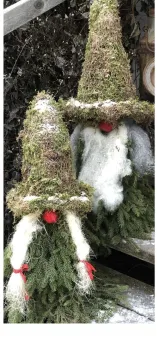 Två dekorativa tomtar med mossmössor och röda näsor, stående i granris med lätt snö mot mörk bakgrund..