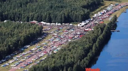 Flygvy över en stor bilträff med många parkerade bilar och besökare längs en väg bredvid en sjö och skog..