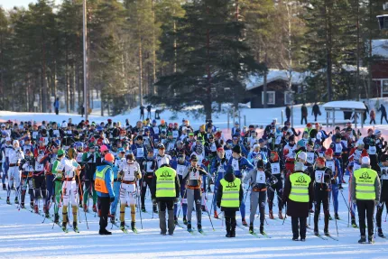 Stor grupp längdskidåkare står samlade vid start på snö, med funktionärer i reflexvästar och skog i bakgrunden..