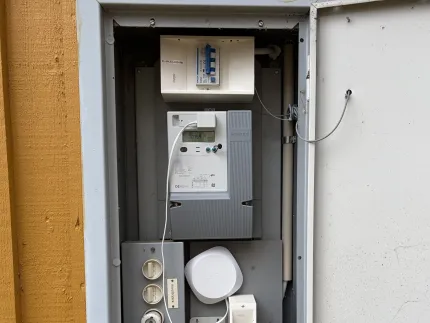 Öppet elskåp på yttervägg med elmätare, säkringar, kablar och en liten vit enhet monterad inuti..