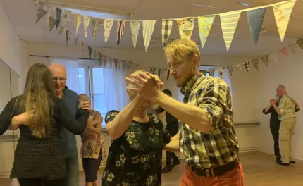 Flera par dansar i en ljus lokal med trägoIv och färgglada vimplar i taket, en man i rutig skjorta i förgrunden..
