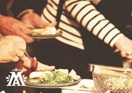 Närbild av händer som serverar mat vid en buffé, med tallrikar, sallad, bestick och en skål på bordet..