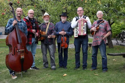 Sex män står på gräs i en trädgård med fruktträd och håller kontrabas, klarinett, gitarr, fiol och dragspel..
