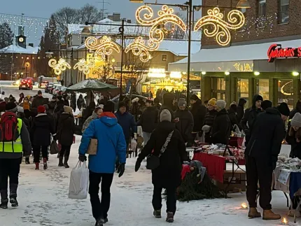 Snötäckt stadsgata med folkmassa, marknadsstånd och varma julbelysningar utanför butiker i skymning..