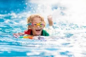 Ett leende barn med simglasögon simmar i en blå pool, med vattenskvätt och soligt ljus runtom..