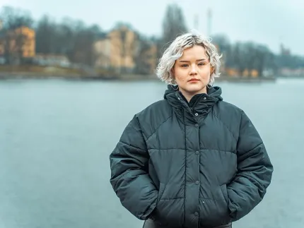 En person i mörk vadderad vinterjacka står vid en stilla sjö, med suddiga byggnader och träd i bakgrunden..