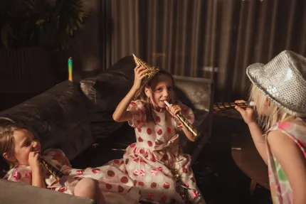 Tre barn i prickiga klänningar firar på en soffa med partyhattar och tutor i ett dämpat vardagsrum..