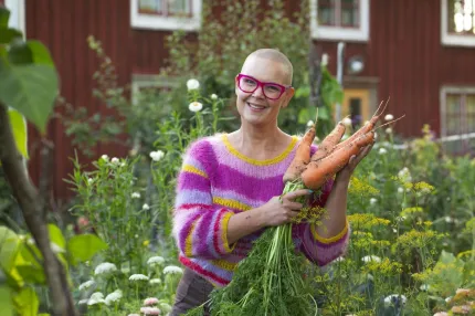 Leende person med rosa glasögon håller en bunt stora morötter i en blommande trädgård framför ett rött trähus..
