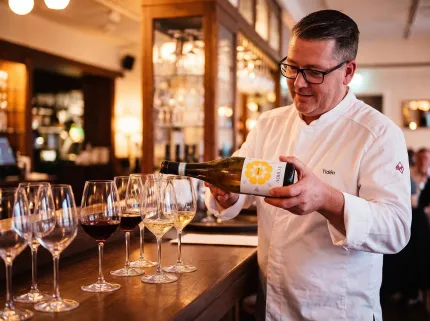 En man i vit kockjacka och glasögon häller upp vin i flera vinglas vid en bardisk i en restaurang..
