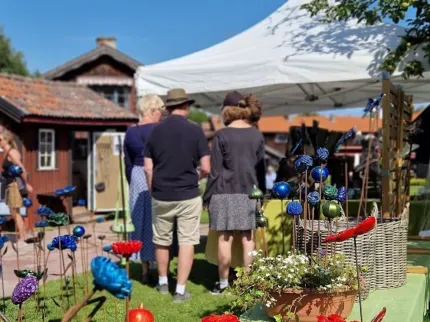 Personer står vid ett marknadsstånd under vitt tält, med färgglada dekorationsblommor och krukväxter i förgrunden.